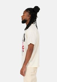 Crème-kleurig T-shirt met een bloemenprint in roze en groen, korte mouwen, ontspannen fit, gecombineerd met lichtbeige broeken.