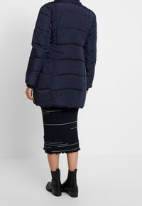 Navy puffer coat with a long silhouette, quilted texture, and high collar. Worn over a black ribbed skirt with white stripes and ankle boots.