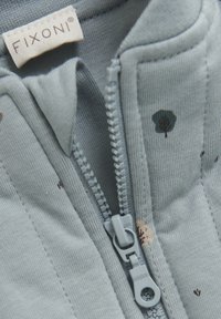 Ljusblå zip-up plagg med grå dragkedja. Har små blommönster i mörka färger, tillverkat av mjukt bomullstyg. Varumärkesetikett synlig.