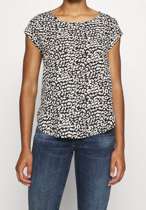 Femme portant un haut à manches courtes imprimé léopard noir et blanc associé à un jean en denim bleu, debout, les bras détendus.