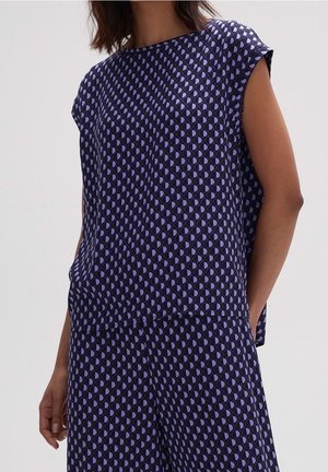 Ärmelloses marineblaues Top und Hose mit sich wiederholendem blauem und hellviolettem geometrischem Halbkreismuster, getragen von einer Person mit der Hand in der Tasche.