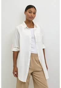 Camisa branca de botão feita de tecido leve, com um bolso no peito e mangas enroladas. Usada sobre uma camisola branca e calças bege.