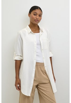 Camicia bianca in lino con colletto, chiusura a bottoni e maniche arrotolate; indossata sopra una canotta bianca e pantaloni beige rilassati.