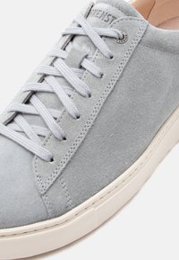 Αθλητικό παπούτσι από ανοιχτό γκρι σουέντ με κορδόνια στο ίδιο χρώμα, λεπτομερείς ραφές και λευκή καουτσούκ σόλα, εμφανιζόμενο σε λευκό φόντο.