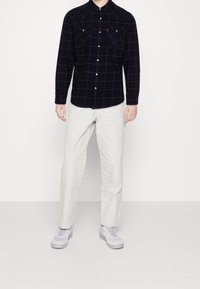 Marinblå rutig långärmad skjorta med knappar, två bröstfickor, ihop med ljusa byxor och grå sneakers.