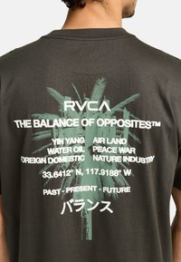 Dunkelgraues T-Shirt mit grünen Palmbaum-Graphics und Text, darunter "RVCA" und "DIE BALANCE DER GEGENSÄTZE", mit weißen Akzenten.
