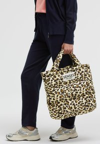 Leopard-print tote bag with a beige base, padded design, and dual handles. "SENSES .THE LABEL" tag on the front. Styled with navy joggers and sneakers.