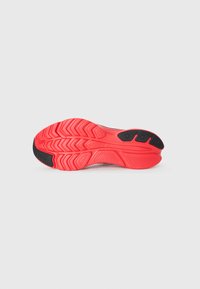 Rød- og sortmønstret gummisål på en Saucony sportsko, set fra neden på en hvid baggrund.
