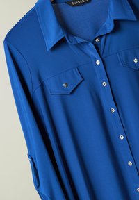 Camicia azzurra con colletto, due tasche frontali e bottoni argentati. Realizzata in un tessuto liscio e leggero con una texture rigata.