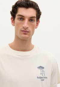 Cremefarvet bomulds t-shirt med et marineblåt broderet design af en strandparasol og stol, med teksten "Scalpers Swim Club".