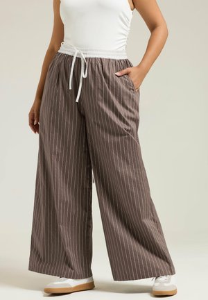 Person iført bredbenede brune bukser med hvide striber, med hvid snøre i taljen og hvide sneakers, stående med den ene hånd i lommen.