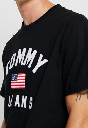 Personne portant un t-shirt noir avec le texte blanc "TOMMY JEANS" et un drapeau américain cousu sur la poitrine, tatouage visible sur le haut du bras.