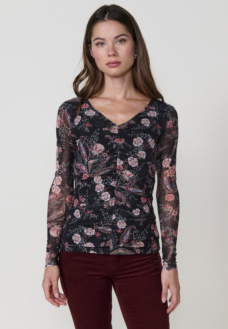 Blusa de manga larga en tejido negro transparente con un patrón floral, que presenta un escote en forma de V y detalles fruncidos en la parte delantera. Combinada con pantalones burdeos oscuros.