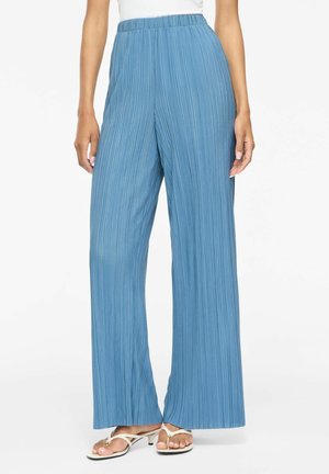 Pantalon plissé bleu taille haute à jambes larges, porté avec des sandales blanches à brides et un haut blanc, présenté de la taille aux pieds sur fond blanc.