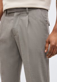 Κοντινό πλάνο ενός ατόμου που φοράει ανοιχτό γκρι παντελόνια προσαρμοσμένης ραφής και λευκό πουκάμισο, εμφανίζοντας τις πλευρικές και εμπρόσθιες τσέπες και το χέρι που ακουμπάει δίπλα στο πόδι.