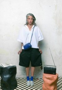 Camicia bianca oversize, pantaloni corti neri, borsa a tracolla blu, scarpe argentate e calze blu, posando davanti a un muro testurizzato e una radio su un supporto in argilla.