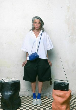 Chemise blanche surdimensionnée, shorts noirs, sac bandoulière bleu, chaussures argentées et chaussettes bleues, posés devant un mur texturé et une radio sur un support en argile.