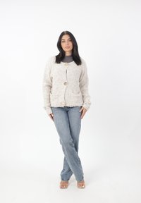 Ljusbeige kofta med fluffig textur, med två framfickor, rund halsringning och stora knappar. Bärs över en grå topp och jeans.