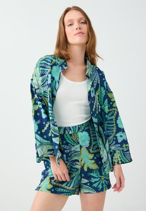 Jeune femme portant un débardeur blanc avec une chemise oversize à motifs bleus et verts et un short assorti, debout devant un fond uni.