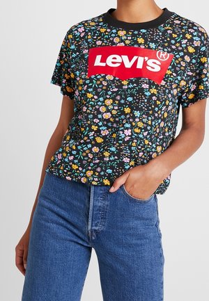 Personne portant un T-shirt Levi's noir à motif floral avec un logo rouge et un jean bleu, une main dans la poche, sur un fond uni.