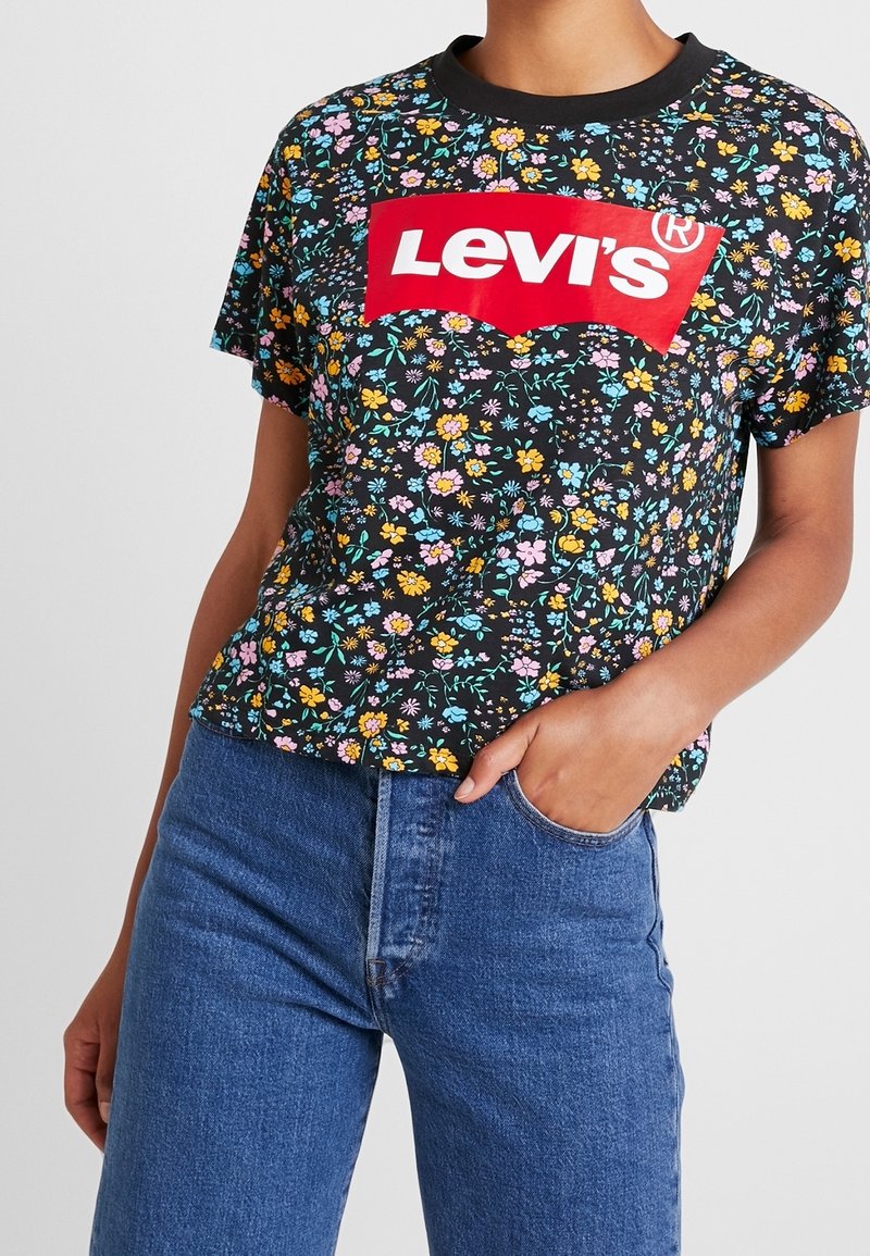 Personne portant un T-shirt Levi's noir à motif floral avec un logo rouge et un jean bleu, une main dans la poche, sur un fond uni.