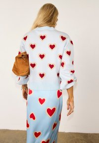 Γυναίκα με μακριά ξανθά μαλλιά που φορά ένα ανοιχτό μπλε πουλόβερ και φούστα με κόκκινα καρδιές, κρατώντας μια καφέ σουέντ τσάντα κάτω από το μπράτσο της.