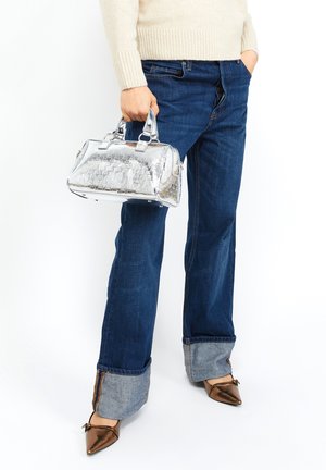Srebrna torebka z lakierowanej skóry o strukturalnym kształcie, z uchwytami do noszenia i teksturowaną powierzchnią. Połączona z ciemnoniebieskimi dżinsami i brązowymi czółenkami w szpic.