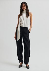 Hihaton pusero vaaleassa beige-kankaassa, jossa korkea kaulus ja pitkä huiviyksityiskohta; yhdistettynä löysiin mustiin housuihin ja mustiin kärjellisiin kenkiin.