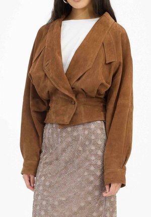 Donna che indossa una giacca oversize in camoscio marrone con grandi risvolti sopra una maglia bianca e una gonna taupe con perline a motivo a conchiglia.