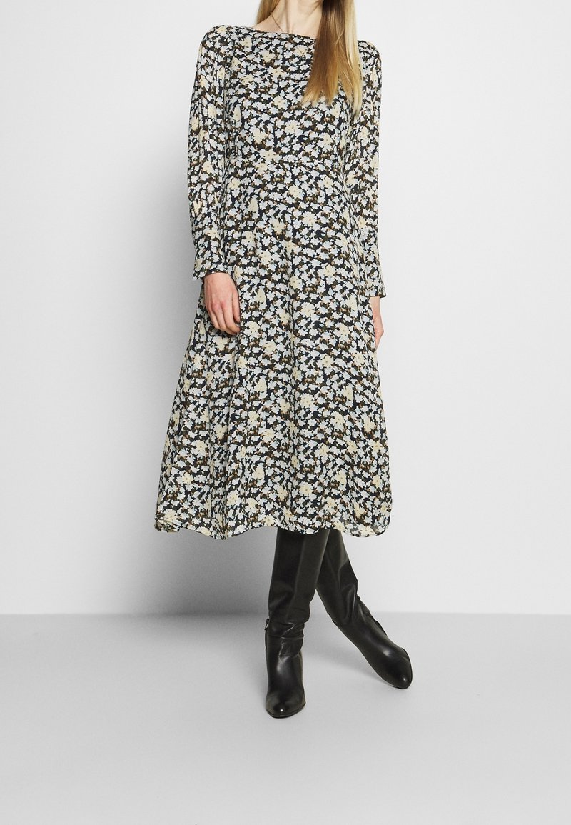 Vestido largo de flores con mangas largas y cuello redondo, con un fondo negro y flores blancas y beige. Llevado con botas negras hasta la rodilla.