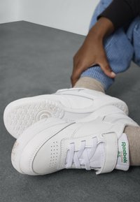 Bjele Reebok tenisice s kožnim gornjim dijelom, čipkastim i čičak zatvaranjem, teksturiranim potplatom i zelenim logotipom.