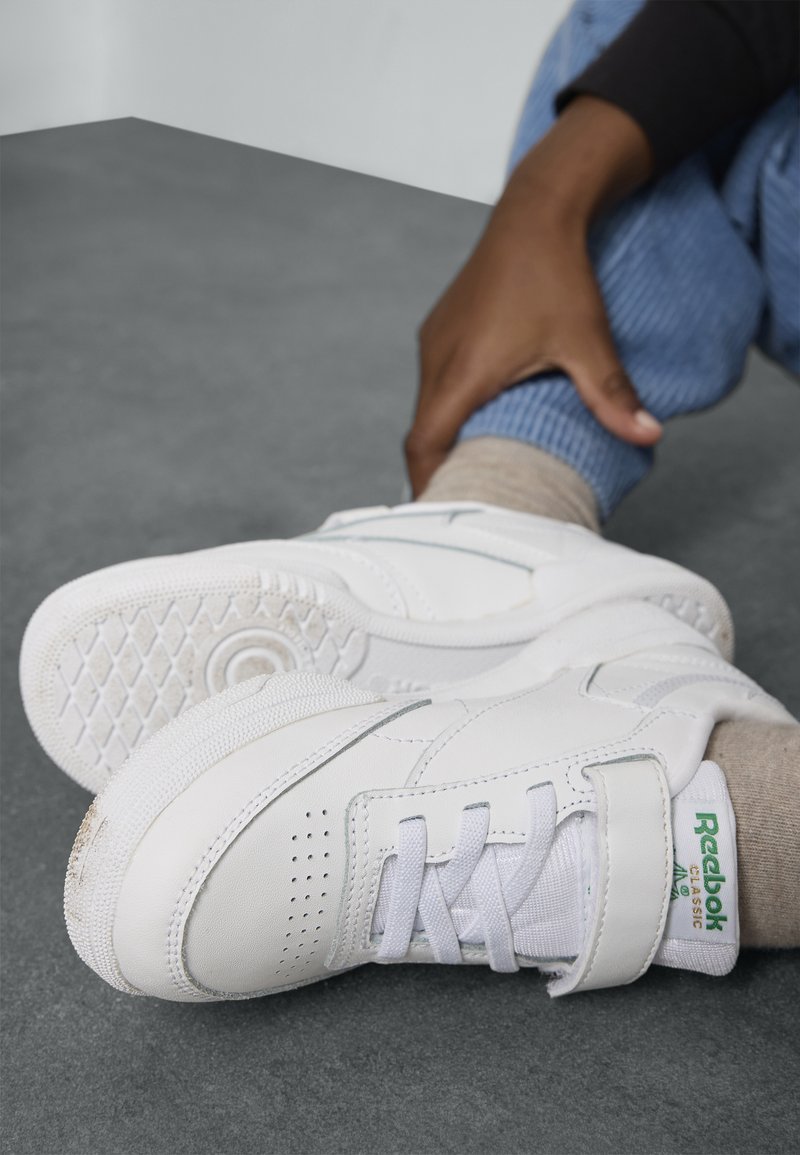 Bjele Reebok tenisice s kožnim gornjim dijelom, čipkastim i čičak zatvaranjem, teksturiranim potplatom i zelenim logotipom.