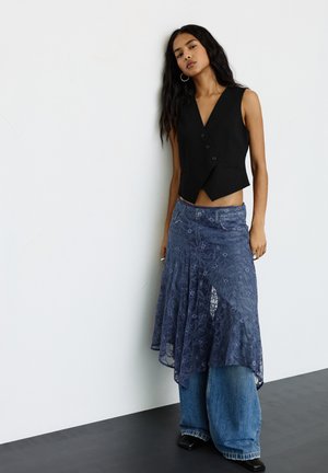 Egy ujjatlan fekete mellényben, kék csipkés szoknyában, széles szárú farmernadrágban és fekete hegyes orrú cipőben álló nő egy fehér fal előtt.