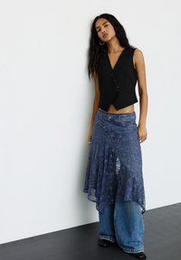 Egy ujjatlan fekete mellényben, kék csipkés szoknyában, széles szárú farmernadrágban és fekete hegyes orrú cipőben álló nő egy fehér fal előtt.