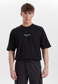 Zwart katoenen T-shirt met korte mouwen en een ronde halslijn, voorzien van een wit bedrukt logo op de borst. Soepele textuur, losse pasvorm.