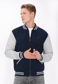 Marineblå og grå varsity jakke med lynlås, stripede ribbede krave og manchetter samt broderede detaljer på fronten.