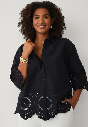 Lächelnde Frau mit lockigem Haar, die eine schwarze Bluse mit kreisförmigen Ausschnitten und weiße Hosen trägt, eine Hand in der Tasche, goldene Armbänder.