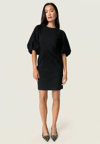Schwarzes kurzes Kleid mit Puffärmeln und Rundhalsausschnitt. Glatter Stoff, figurbetonte Silhouette und knielanges Design. Ein Paar schwarz-weiß gestreifte High Heels.