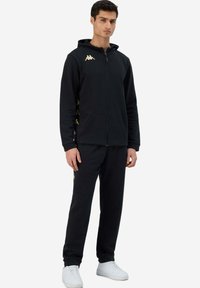 Felpa nera con zip e pantaloni abbinati, caratterizzati da loghi dorati sul petto e sul lato. Il tessuto è liscio; il design include una tasca frontale.