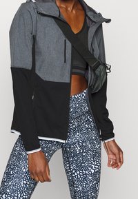 Veste de sport grise et noire, avec une fermeture éclair à l'avant et des poignets élastiques ; associée à des leggings à motifs bleu marine et blancs. Sac d'accessoires inclus.