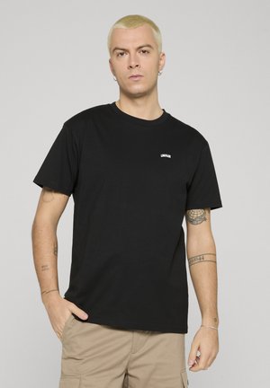 Hombre rubio con pendientes y tatuajes que lleva una camiseta negra con el logo "UNFAIR" y pantalones cargo beige, de pie frente a un fondo liso.