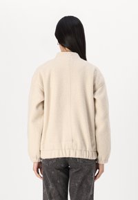 Veste en polaire couleur crème avec un col haut, un ourlet élastique et un tissu texturé. La vue de dos montre une coupe décontractée et des coutures.