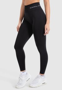 Leggings neri realizzati in materiale elasticizzato presentano un ampio cinturino con il marchio "SMILODON". Indossati con sneakers bianche e calze.