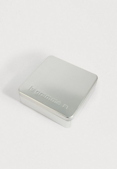 Pequena caixa de metal prateada com bordas arredondadas, em relevo com as palavras "le gramme" e um quadrado na tampa, colocada sobre uma superfície branca.