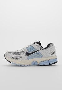 Zapatilla de running Nike en blanco y azul claro con swoosh negro, paneles de malla y suela texturizada sobre un fondo blanco liso.