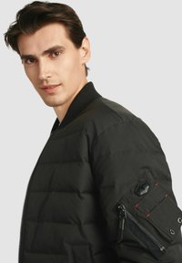 Czarna kurtka puchowa z pikowanym wzorem, wywijanym kołnierzem i kieszenią na zamek błyskawiczny na lewym rękawie, z czerwonymi akcentami i błyszczącym zamkiem.