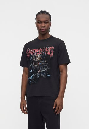 Młody mężczyzna z warkoczami, ubrany w czarną koszulkę z nadrukiem z czerwonym napisem i projektem mrocznego wojownika fantasy na białym tle.