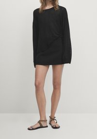 Zwarte lange mouw jurk shirt met een gedraaide knoopdetail, gecombineerd met platte zwarte sandalen met een metalen accent en bruine banden.