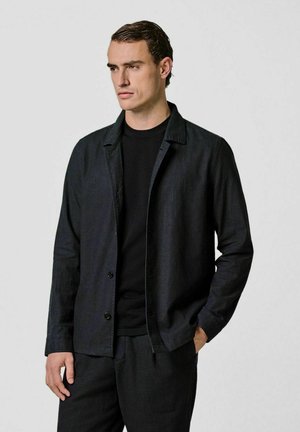 Chaqueta de color oscuro hecha de tela con una textura suave, con cuello, cierre de botones y un ajuste cómodo, combinada con una camisa negra.