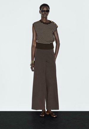 Gestreiftes Strickoberteil in Braun und Creme mit Schulterdetails, kombiniert mit weiten braunen Hosen. Getragen mit schwarzen Sonnenbrillen und flachen Sandalen.